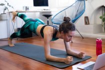 Femme caucasienne passant du temps à la maison, portant des vêtements de sport, faisant de l'exercice sur un tapis, rejoignant un cours de yoga en ligne, utilisant son ordinateur portable. Distance sociale et isolement personnel en quarantaine. — Photo de stock