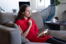 Caucasian woman spending time at home, wearing a pink dress, sitting on a sofa and reading a book. Social distancing and self isolation in quarantine lockdown. — Stock Photo