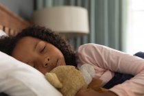 Афроамериканська дівчинка спить у спальні свого батька і обіймає плюшевого ведмедика, під час соціального дистанціювання вдома під час карантину.. — стокове фото