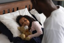 African American man waking up his daughter, sleeping in his bed and embracing a teddy bear, during social distancing at home during quarantine lockdown. — Stock Photo