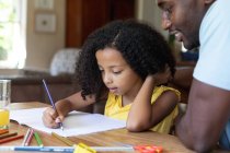 Afroamerikanerin in gelber Bluse, soziale Distanzierung zu Hause während der Quarantäne, am Tisch sitzend und Bilder mit ihrem Vater zeichnend. — Stockfoto