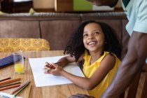 Menina afro-americana vestindo uma blusa amarela, distanciamento social em casa durante o confinamento de quarentena, sentada ao lado de uma mesa e desenhando fotos com seu pai . — Fotografia de Stock