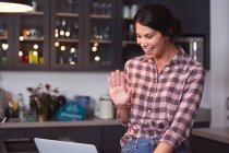 Mixed race woman spending time at home self isolating and social distancing in quarantine lockdown during coronavirus covid 19 epidemic, smiling, waving, using laptop in kitchen. — Stock Photo
