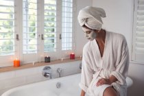Mixed race woman spending time at home, sitting on bathtub with face mask on running bath in bathroom. Self isolating and social distancing in quarantine lockdown during coronavirus covid 19 epidemic. — Stock Photo
