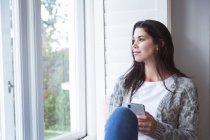 Змішана жінка проводить час вдома, сидячи на віконному сидінні, тримаючи смартфон у вітальні. Самоізоляція та соціальне дистанціювання в карантинному блокуванні під час епідемії коронавірусу 19 . — стокове фото