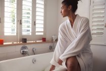Жінка змішаної раси проводить час вдома, сидячи на ванні, що працює у ванній. Самоізоляція та соціальне дистанціювання в карантинному блокуванні під час епідемії коронавірусу 19 . — стокове фото