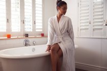 Смешанная расовая женщина проводит время дома самоизоляция и социальное дистанцирование в карантинной изоляции во время эпидемии коронавируса ковид 19, сидя на ванне работает ванна в ванной комнате. — стоковое фото