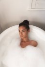 Смешанная расовая женщина проводит время дома самоизоляция и социальное дистанцирование в карантинной изоляции во время эпидемии коронавируса ковид 19, лежа в ванне, покрытой пеной расслабляющий в ванной комнате. — стоковое фото