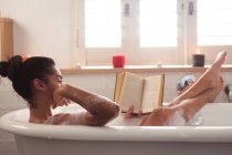 Mixed race woman spending time at home self isolating and social distancing in quarantine lockdown during coronavirus covid 19 epidemic, lying in bathtub reading a book relaxing in bathroom. — Stock Photo
