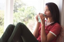 Gemischte Rassenfrau, die während der Coronavirus-Covid 19-Epidemie Zeit zu Hause mit Selbstisolierung und sozialer Distanzierung in Quarantäne zubringt, sitzt auf Fensterbank und trinkt Kaffee im Wohnzimmer. — Stockfoto