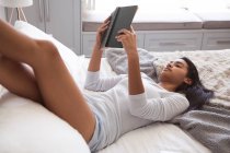 Mixed race woman spending time at home self isolating and social distancing in quarantine lockdown during coronavirus covid 19 epidemic, lying on bed reading a book in bedroom. — Stock Photo