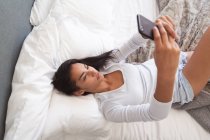 Mixed race woman spending time at home, lying on bed taking selfies with her smartphone in bedroom. Self isolating and social distancing in quarantine lockdown during coronavirus covid 19 epidemic. — Stock Photo