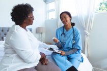 Seniorin mit gemischter Rasse verbringt Zeit zu Hause, wird von einer Krankenschwester mit gemischter Rasse besucht, sitzt auf einem Bett und redet — Stockfoto
