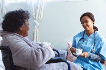 Старшая женщина смешанной расы проводит время дома, сидит в инвалидном кресле, ее посещает медсестра смешанной расы, держит чашки и разговаривает — стоковое фото