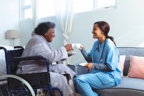 Старшая женщина смешанной расы проводит время дома, сидит на инвалидной коляске, ее посещает медсестра смешанной расы, держит чашки и разговаривает — стоковое фото