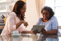 Mujer de raza mixta mayor que pasa tiempo en casa con su hija, distanciamiento social y aislamiento en cuarentena, usando una tableta - foto de stock