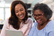 Mujer de raza mixta mayor que pasa tiempo en casa con su hija, distanciamiento social y aislamiento en cuarentena, usando una tableta - foto de stock