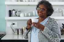 Senior donna razza mista godendo il suo tempo a casa, distanza sociale e auto isolamento in quarantena isolamento, in piedi nella sua cucina, tenendo una tazza e sorridente — Foto stock
