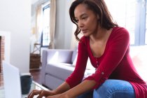 Mulher de raça mista aproveitando seu tempo em casa, distanciamento social e auto-isolamento em quarentena, sentada em um sofá, usando um laptop — Fotografia de Stock