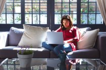 Gemischte Rassenfrau genießt ihre Zeit zu Hause, soziale Distanzierung und Selbstisolierung in Quarantäne, sitzt auf einem Sofa und benutzt einen Laptop — Stockfoto