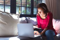 Mulher de raça mista aproveitando seu tempo em casa, distanciamento social e auto-isolamento em quarentena, sentada em um sofá, usando um laptop — Fotografia de Stock