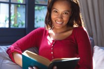 Porträt einer Frau mit gemischter Rasse, die ihre Zeit zu Hause genießt, soziale Distanzierung und Selbstisolierung in Quarantäne, auf einem Sofa sitzend, ein Buch in der Hand, in die Kamera schauend und lächelnd — Stockfoto