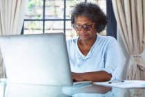 Seniorin mit gemischter Rasse genießt ihre Zeit zu Hause, soziale Distanzierung und Selbstisolierung in Quarantäne, sitzt an einem Tisch und benutzt einen Laptop — Stockfoto
