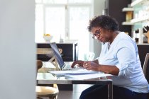 Mujer de raza mixta mayor disfrutando de su tiempo en casa, distanciamiento social y aislamiento en cuarentena, sentada en una mesa, usando una computadora portátil, haciendo papeleo - foto de stock