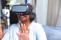Senior mixed race woman enjoying her time at home, social distancing and self isolation in quarantine lockdown, sitting on a sofa, wearing vr goggles and touching virtual reality screen — Stock Photo