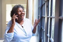Seniorin mit gemischter Rasse genießt ihre Zeit zu Hause, soziale Distanzierung und Selbstisolierung in Quarantäne, schaut aus dem Fenster, spricht auf dem Smartphone — Stockfoto