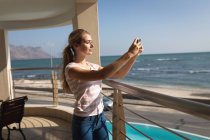 Donna caucasica in piedi su un balcone, che tiene uno smartphone e si fa un selfie. Distanziamento sociale e autoisolamento in quarantena. — Foto stock