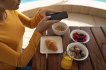 Mujer caucásica sentada junto a una mesa, tomando una foto del desayuno con su smartphone. Distanciamiento social y autoaislamiento en cuarentena. - foto de stock