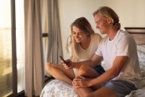 Kaukasisches Paar, das zusammen im Bett sitzt und ein Smartphone benutzt. Soziale Distanzierung und Selbstisolierung in Quarantäne. — Stockfoto