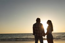 Кавказька пара стоїть на пляжі під час заходу сонця, тримаючись за руки і дивлячись один на одного. — стокове фото