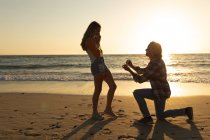 Couple caucasien profitant de leur temps sur une plage pendant le coucher du soleil, un homme s'agenouille et demande en mariage à une femme — Photo de stock