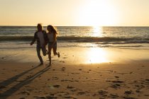 Kaukasisches Paar im Urlaub am Strand bei Sonnenuntergang, Händchen haltend, während sich die untergehende Sonne im Meer und Sand hinter ihnen spiegelt — Stockfoto