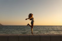 Mujer caucásica de vacaciones disfrutando de su tiempo en un paseo marítimo durante una puesta de sol, saltando y extendiendo sus brazos, el sol poniéndose sobre el mar en el fondo - foto de stock