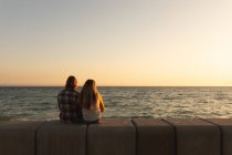 Vista trasera de la pareja caucásica sentados juntos en un paseo marítimo al atardecer, mirando al mar. Romántica pareja de vacaciones de playa - foto de stock
