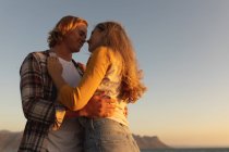 Kaukasisches Paar, das bei Sonnenuntergang auf einer Promenade steht, sich umarmt und küsst. Romantisches Paar Urlaub am Meer — Stockfoto