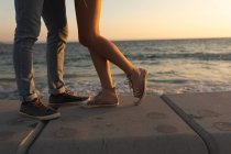 Section basse du couple debout sur une promenade au bord de la mer au coucher du soleil, face à face et embrassant ou embrassant. Romantique couple de vacances à la mer — Photo de stock