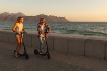 Casal caucasiano montando e-scooters em um passeio marítimo durante o pôr do sol, olhando uns para os outros, relaxando durante um feriado ativo na praia à beira-mar — Fotografia de Stock