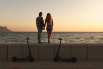 Rückansicht eines kaukasischen Paares, das bei Sonnenuntergang mit geparkten E-Scootern auf einer Strandpromenade am Meer steht, sich an den Händen hält und auf das Meer blickt. Paar im romantischen Strandurlaub am Meer — Stockfoto