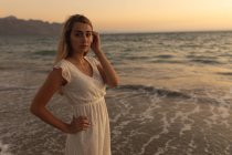 Портрет кавказької жінки, одягненої в білу сукню, що стоїть на пляжі під час заходу сонця, дивиться на камеру, відпочиває під час активного берегового відпочинку. — стокове фото
