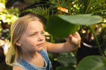 Une fille caucasienne aux longs cheveux blonds profitant du temps dans un jardin ensoleillé, explorant, touchant des feuilles de plantes et les regardant — Photo de stock