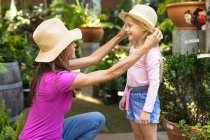 Una mujer caucásica y su hija disfrutando del tiempo juntos en un jardín soleado, mujer arrodillada y poniéndose un sombrero en la cabeza de sus hijas, sonriéndose - foto de stock