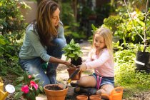 A Caucasian woman and her daughter enjoying time together in a sunny garden, planting a seedling in a plant pot — Stock Photo