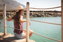 Девушка-подросток в соломенной шляпе, наслаждающаяся прогулкой, в солнечный день, сидя и опираясь на барьер, отворачиваясь и улыбаясь — стоковое фото