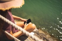Por encima de la vista del hombro de una adolescente caucásica, con un sombrero de paja, disfrutando de su tiempo en un paseo marítimo, en un día soleado, sentado y usando un teléfono inteligente - foto de stock