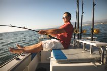Un hombre caucásico disfrutando de su tiempo de vacaciones en el sol junto a la costa, sentado en un barco, sosteniendo una caña de pescar - foto de stock