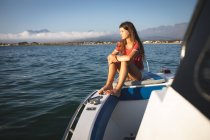 Una adolescente caucásica disfrutando de su tiempo de vacaciones en el sol junto a la costa, sentada en un barco, relajándose, mirando hacia otro lado - foto de stock